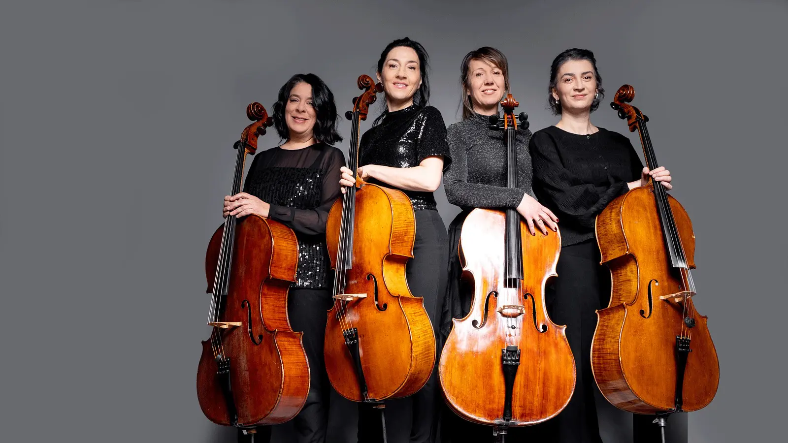 Gruppenporträt von vier Cellistinnen und Cellisten vor grauem Hintergrund. Die Musikerinnen und Musiker stehen nebeneinander in dunkler Konzertkleidung und halten ihre Violoncelli vor sich.