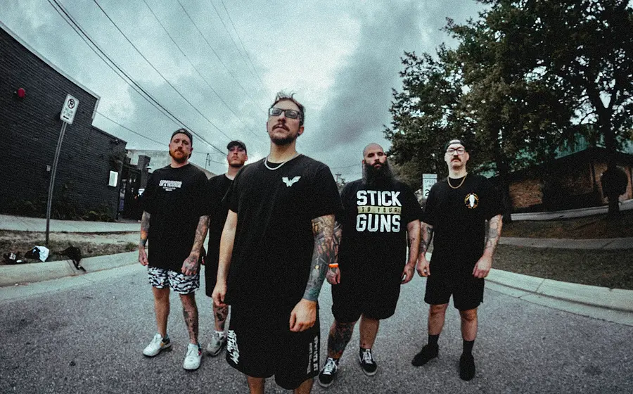 Bandfoto von Fight From Within mit fünf Bandmitgliedern in schwarzer Freizeitkleidung auf einer Straße vor Industriegebäuden, darunter tätowierte Personen mit Brillen, Kappen und langem Bart, aufgenommen mit leichter Weitwinkelperspektive vor bewölktem Himmel