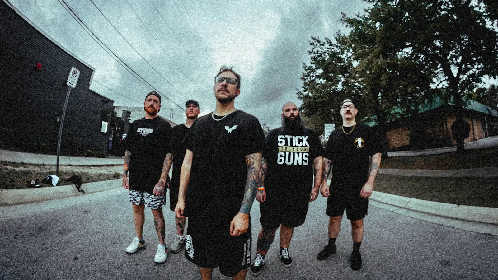 Bandfoto von Fight From Within mit fünf Bandmitgliedern in schwarzer Freizeitkleidung auf einer Straße vor Industriegebäuden, darunter tätowierte Personen mit Brillen, Kappen und langem Bart, aufgenommen mit leichter Weitwinkelperspektive vor bewölktem Himmel