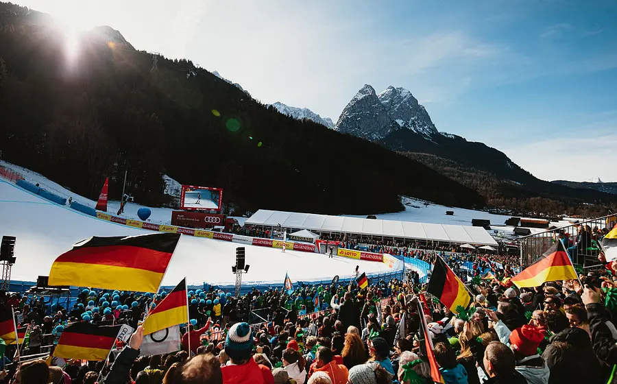 Foto vom Ski-Weltcup in Garmisch-Partenkirchen mit dicht besetzten Zuschauertribünen entlang der Kandahar-Abfahrt, zahlreichen Deutschlandfahnen im Publikum sowie verschneiter Rennstrecke vor Bergkulisse im Sonnenlicht