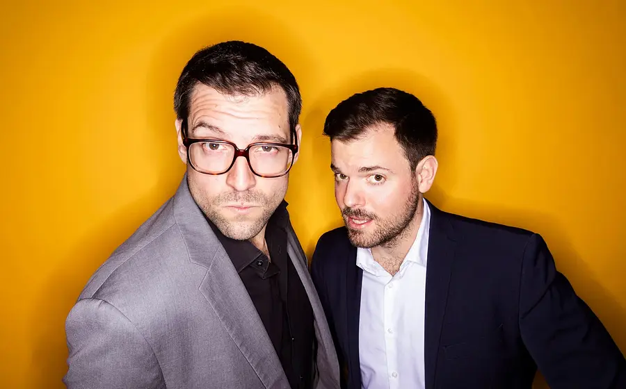 Pressefoto von Flo und Wisch in grauem und dunkelblauem Sakko vor leuchtend orangem Hintergrund, eine Person mit großer Brille im Vordergrund, die andere leicht nach vorne gebeugt daneben.