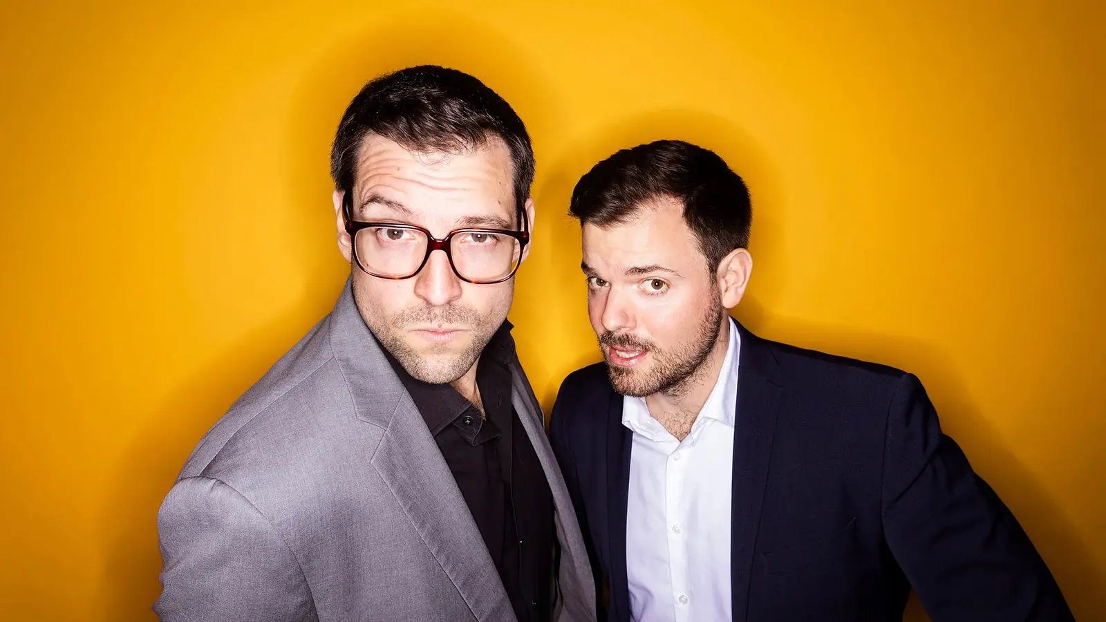 Pressefoto von Flo und Wisch in grauem und dunkelblauem Sakko vor leuchtend orangem Hintergrund, eine Person mit großer Brille im Vordergrund, die andere leicht nach vorne gebeugt daneben.