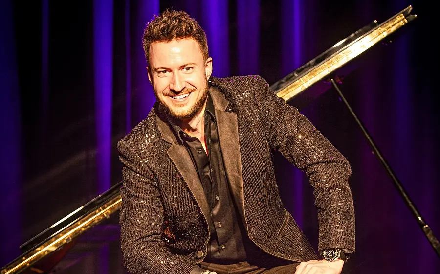 Florian Wagner in einem schwarz glitzernden Jakett sitzt auf einem Konzertflügel und lacht in die Kamera, vor einem violetten Vorhang