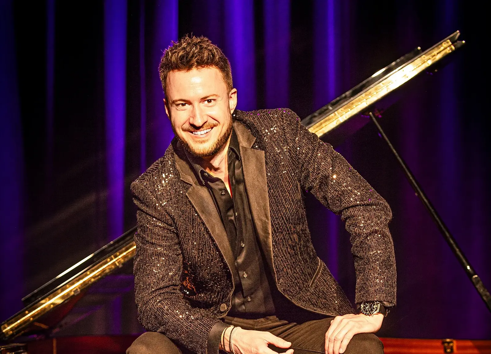 Florian Wagner in einem schwarz glitzernden Jakett sitzt auf einem Konzertflügel und lacht in die Kamera, vor einem violetten Vorhang