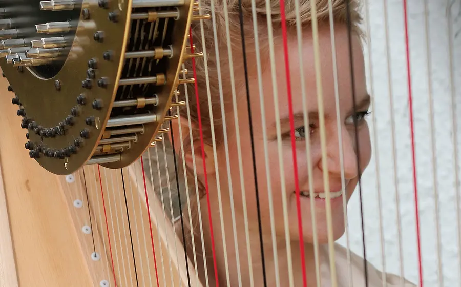 Nahaufnahme einer Harfe mit sichtbaren Saiten und Mechaniken. Hinter den Saiten ist das Gesicht einer Person zu sehen, während die Aufnahme den Blick durch das Instrument richtet.