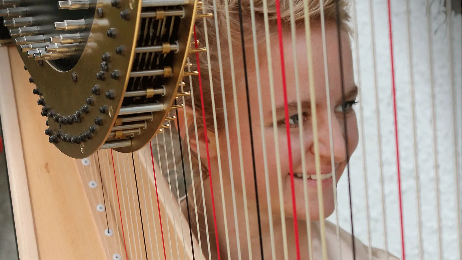 Nahaufnahme einer Harfe mit sichtbaren Saiten und Mechaniken. Hinter den Saiten ist das Gesicht einer Person zu sehen, während die Aufnahme den Blick durch das Instrument richtet.