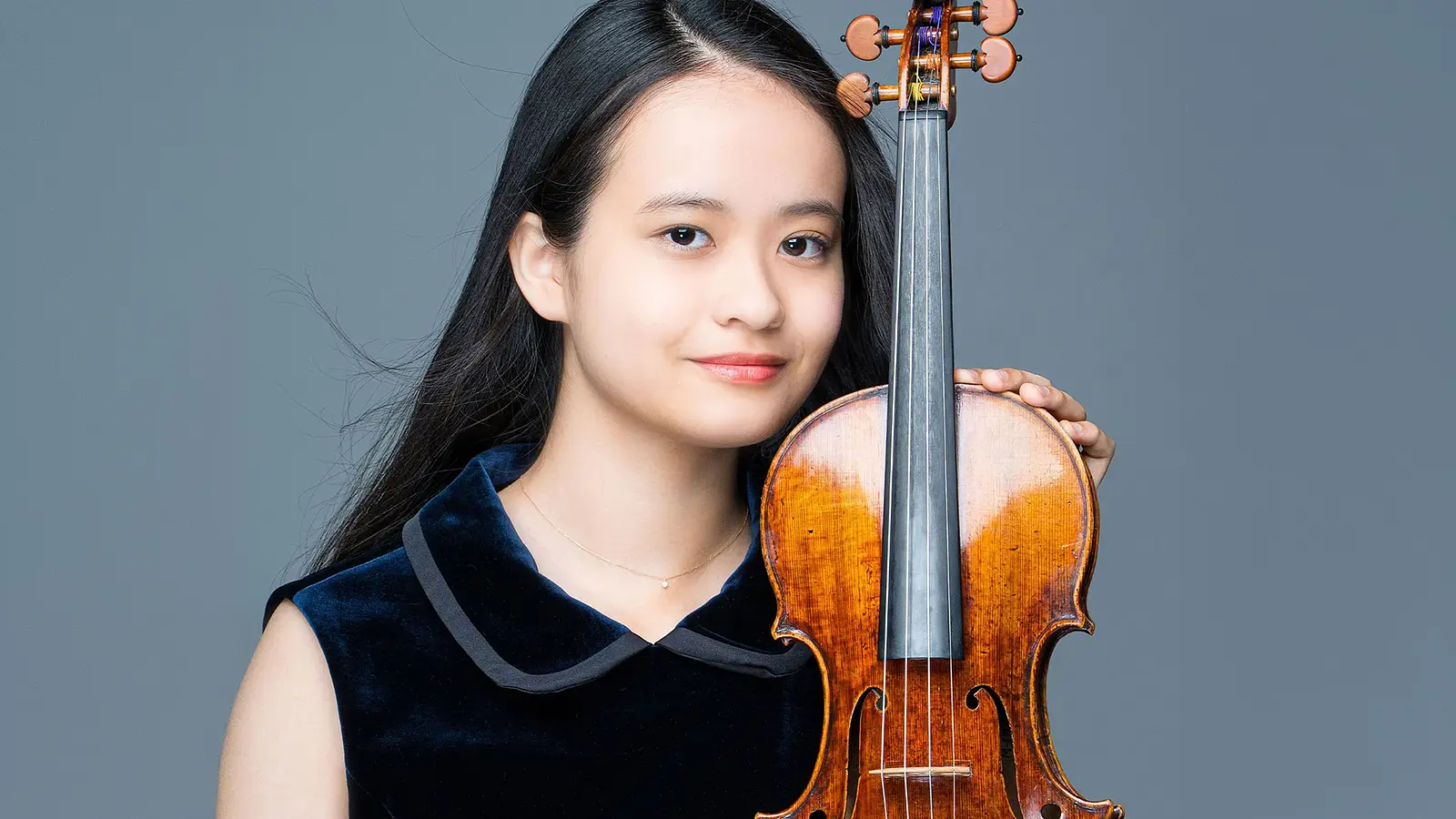 Porträt einer Person mit Violine vor grauem Hintergrund; die Violine wird aufrecht gehalten und steht im Vordergrund des Bildes.