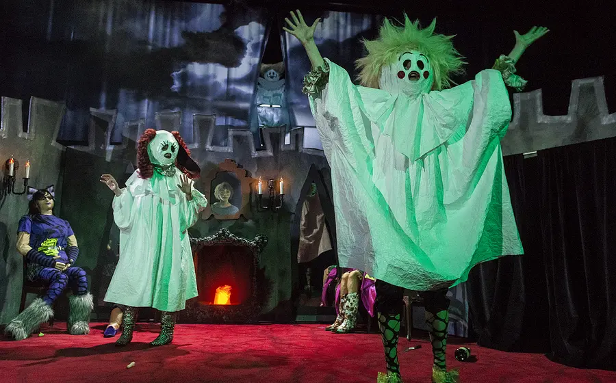 Halloweenartig inszenierte Theaterszene mit mehreren Darstellenden in Geisterkostümen vor einer gruseligen Kulisse mit Schlossmauern, Kerzenleuchtern und Kamin. Eine Figur mit wirrem Haar und ausgebreiteten Armen steht im Vordergrund, während weitere kostümierte Figuren auf der Bühne verteilt sind.