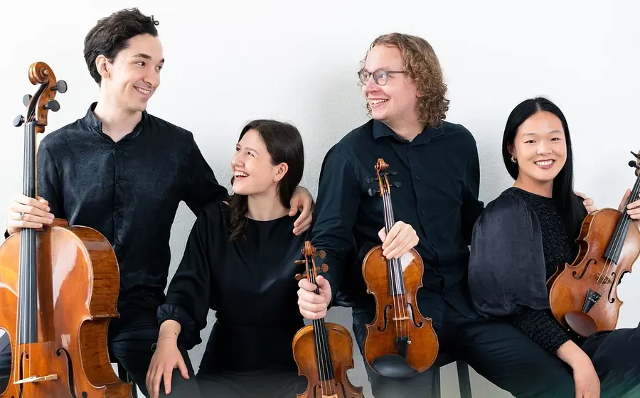 Vier schwarz gekleidete Musiker*innen mit Instrumenten sitzend lachend auf Hockern vor hellem Hintergrund