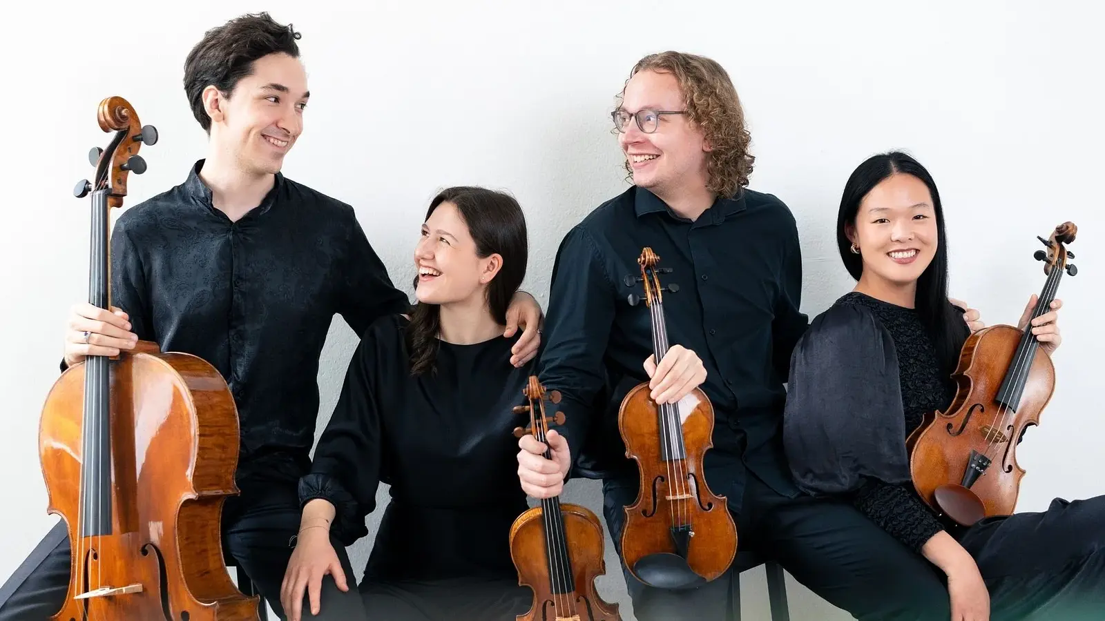 Vier schwarz gekleidete Musiker*innen mit Instrumenten sitzend lachend auf Hockern vor hellem Hintergrund