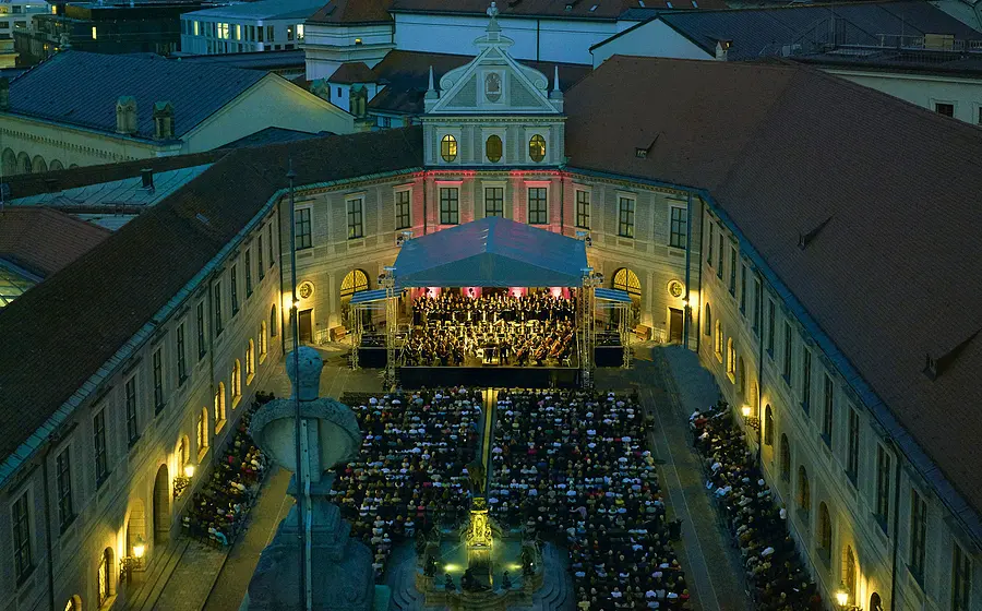 Luftaufnahme eines Open-Air-Konzerts in einem historischen Innenhof bei Abenddämmerung. Vor einer überdachten Bühne mit Orchester und Chor sitzt ein großes Publikum, umgeben von beleuchteten Gebäudefassaden und roter Dachlandschaft.