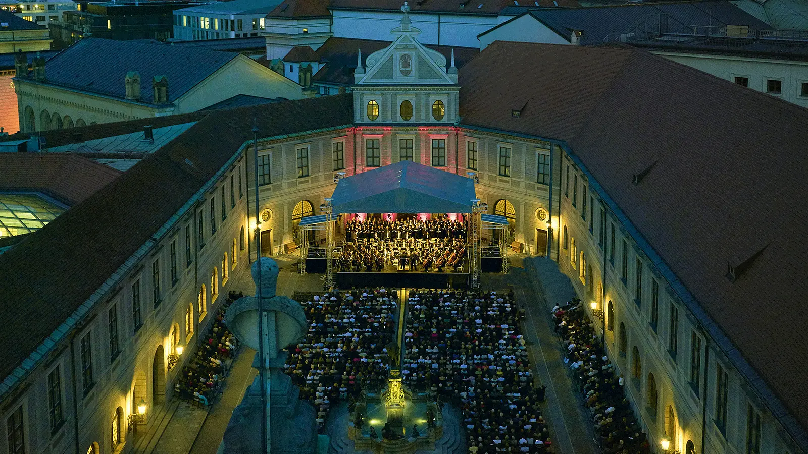 Luftaufnahme eines Open-Air-Konzerts in einem historischen Innenhof bei Abenddämmerung. Vor einer überdachten Bühne mit Orchester und Chor sitzt ein großes Publikum, umgeben von beleuchteten Gebäudefassaden und roter Dachlandschaft.