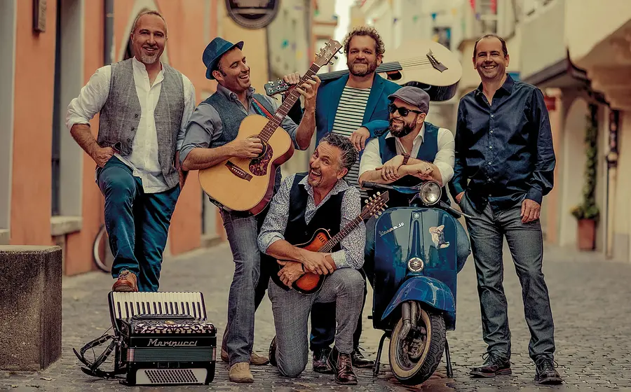 Sechs Musiker mit Gitarren, Akkordeon und Vespa posieren in einer italienisch wirkenden Altstadtgasse.