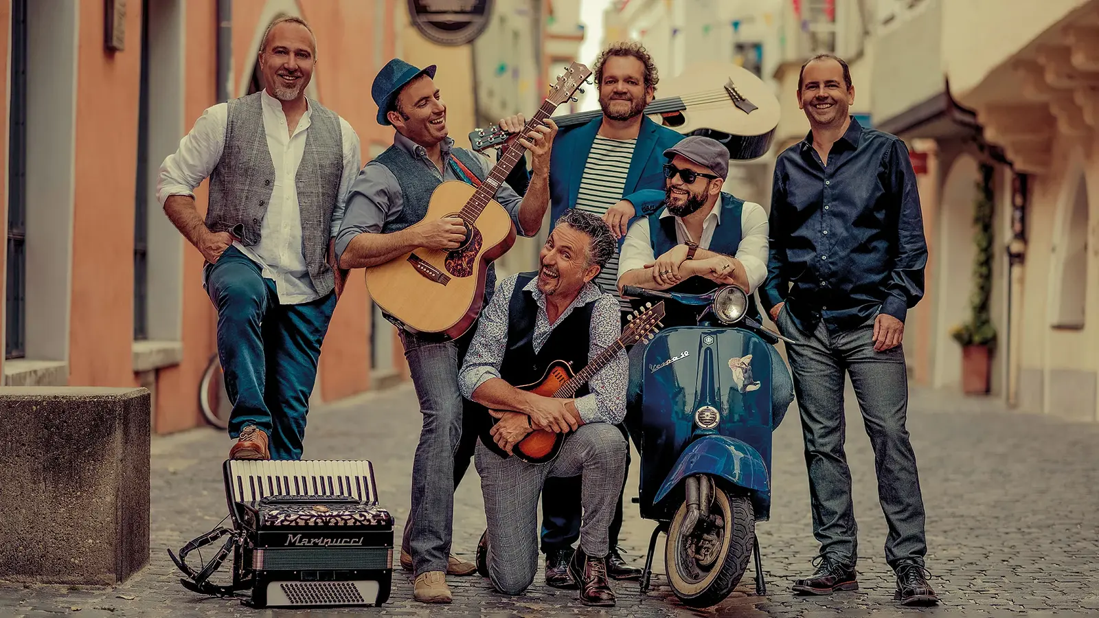 Sechs Musiker mit Gitarren, Akkordeon und Vespa posieren in einer italienisch wirkenden Altstadtgasse.