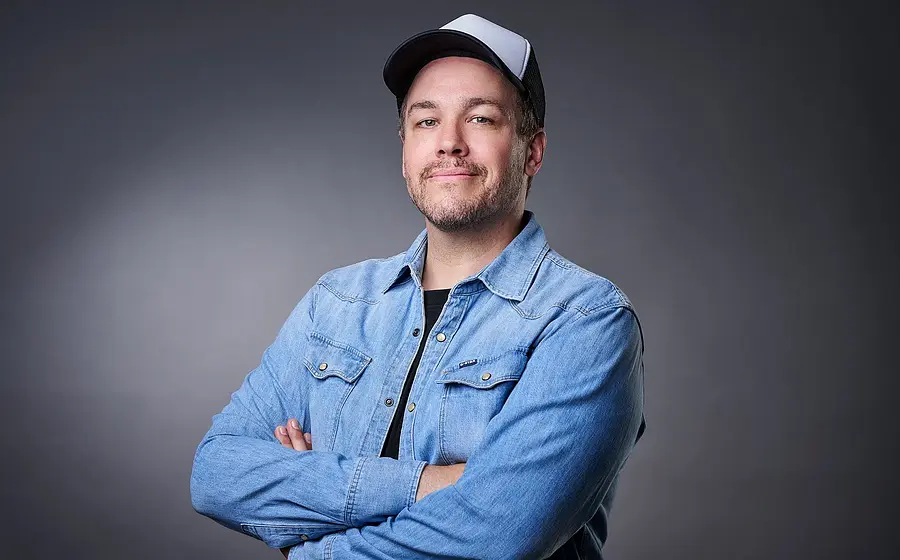 Jan von Weyde steht mit Jeanshemd und weißer Cap vor einem grauen Hintergrund und schaut in die Kamera, während er seine Arme verschränkt.