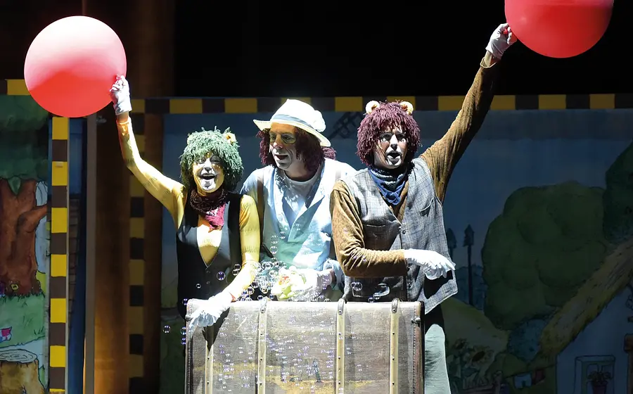 Drei Darsteller in Tierkostümen stehen auf einer Theaterbühne hinter einer Reisekiste und halten rote Luftballons in die Höhe