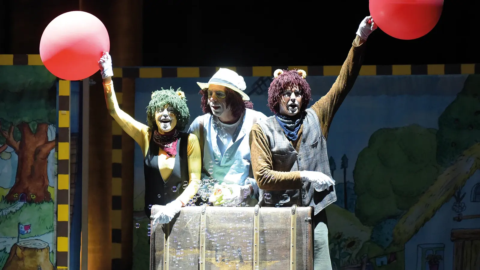 Drei Darsteller in Tierkostümen stehen auf einer Theaterbühne hinter einer Reisekiste und halten rote Luftballons in die Höhe