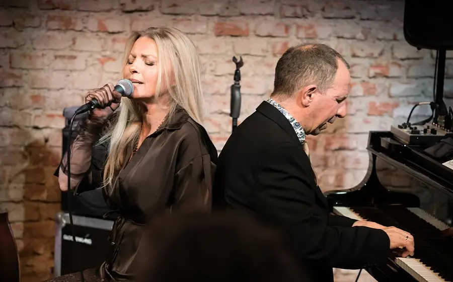 Sängerin Jasmin Bayer mit langem blondem Haar singt in ein Mikrofon, daneben begleitet Davide Roberts sie am schwarzen Flügel. Im Hintergrund eine Backsteinwand, links der Ausschnitt eines Kontrabasses.