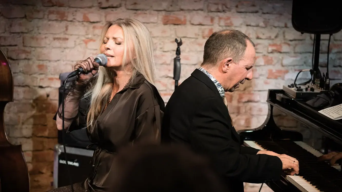 Sängerin Jasmin Bayer mit langem blondem Haar singt in ein Mikrofon, daneben begleitet Davide Roberts sie am schwarzen Flügel. Im Hintergrund eine Backsteinwand, links der Ausschnitt eines Kontrabasses.