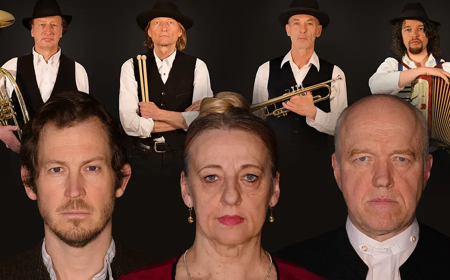 Pressefoto der Musikgruppe Jennerwein: Gruppenporträt vor schwarzem Hintergrund mit mehreren Personen in dunkler, traditionell anmutender Kleidung. Einige halten Blas‑ und Akkordeon‑Instrumente. Drei Personen sind frontal im Vordergrund zu sehen, weitere stehen dahinter. Kontrastreiches Bühnen‑ und Eventmotiv der Band Jennerwein.