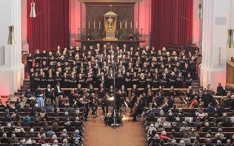 Bühnenfoto des Joseph-Chors München mit großem Chor und Orchester vor Publikum in einer Kirche, vor rotem Vorhang und Altarbereich mit Kerzen, während der Dirigent zentral vor dem Ensemble steht.