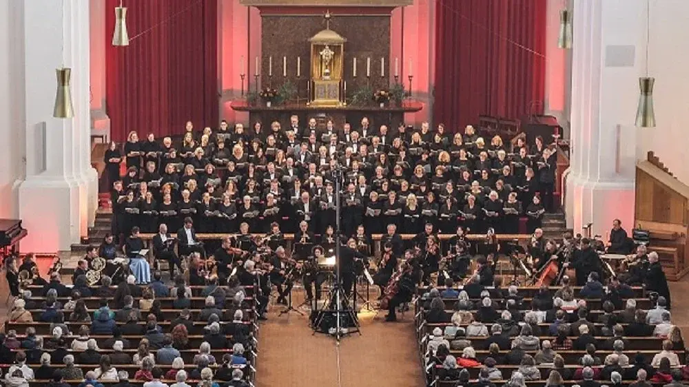 Bühnenfoto des Joseph-Chors München mit großem Chor und Orchester vor Publikum in einer Kirche, vor rotem Vorhang und Altarbereich mit Kerzen, während der Dirigent zentral vor dem Ensemble steht.
