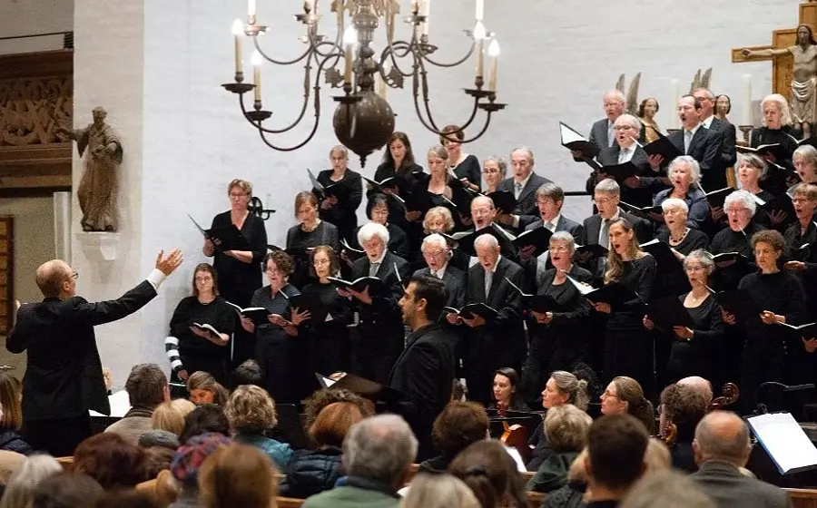 Bühnenfoto eines Chors in schwarzer Konzertkleidung mit Orchester in einer Kirche, vor einem großen Holzkreuz und heller Kirchenwand, mit dirigierender Person im Vordergrund sowie Publikum in den Kirchenbänken