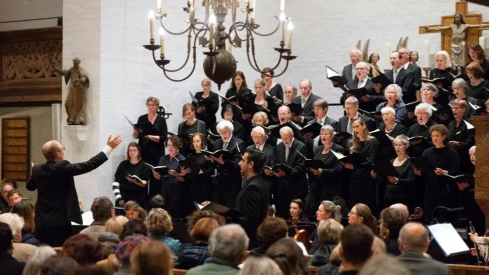 Bühnenfoto eines Chors in schwarzer Konzertkleidung mit Orchester in einer Kirche, vor einem großen Holzkreuz und heller Kirchenwand, mit dirigierender Person im Vordergrund sowie Publikum in den Kirchenbänken