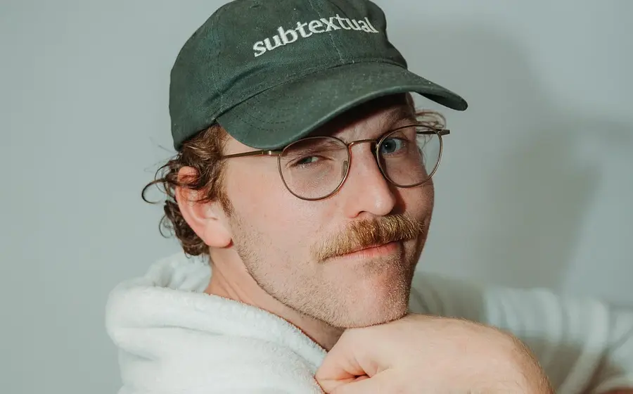 Portrait von Jordan Prince in weißem Pullover mit grüner Kappe blickt lächelnd in die Kamera vor hellem Hintergrund