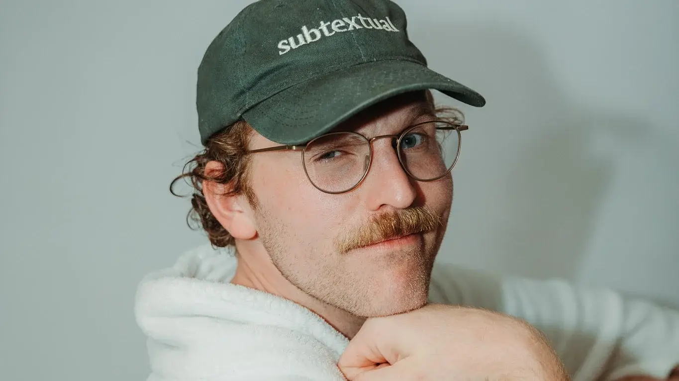 Portrait von Jordan Prince in weißem Pullover mit grüner Kappe blickt lächelnd in die Kamera vor hellem Hintergrund