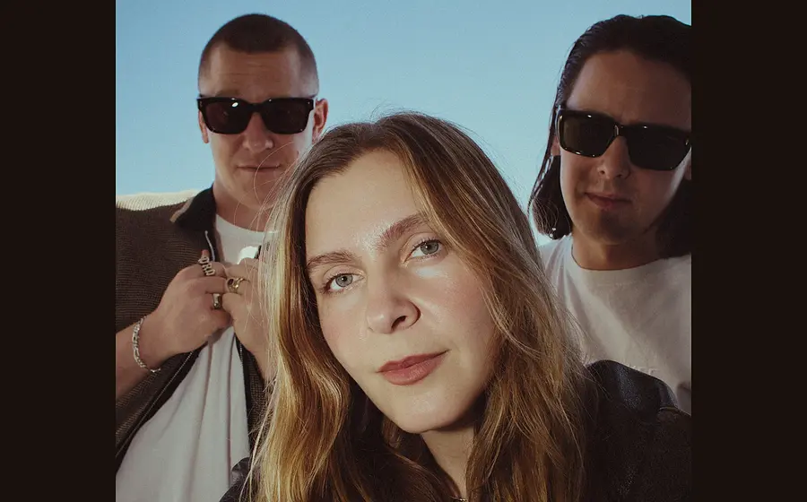 Foto der Band Jungle mit drei Personen, die eng beieinander im Freien vor blauem Himmel stehen; eine Person im Vordergrund mit langen Haaren, die beiden anderen seitlich dahinter in lässiger Kleidung