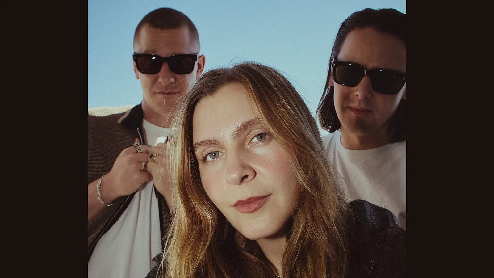 Foto der Band Jungle mit drei Personen, die eng beieinander im Freien vor blauem Himmel stehen; eine Person im Vordergrund mit langen Haaren, die beiden anderen seitlich dahinter in lässiger Kleidung