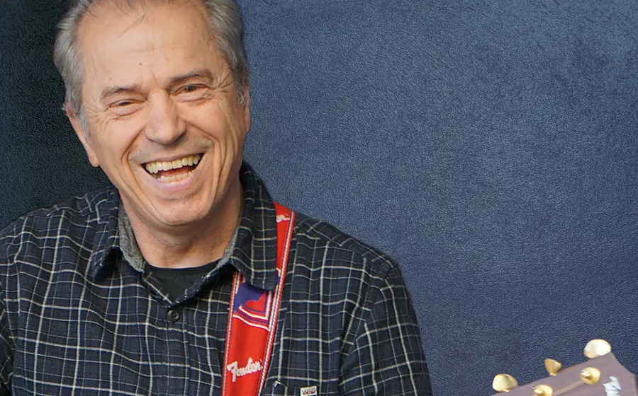 Günther Sigl in einem karierten hemd lachend mit Gitarre vor blau marmoriertem Hintergrund