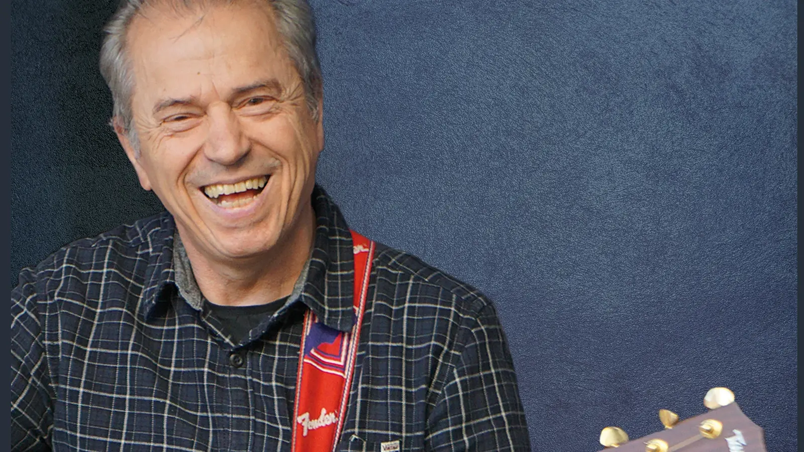Günther Sigl in einem karierten hemd lachend mit Gitarre vor blau marmoriertem Hintergrund