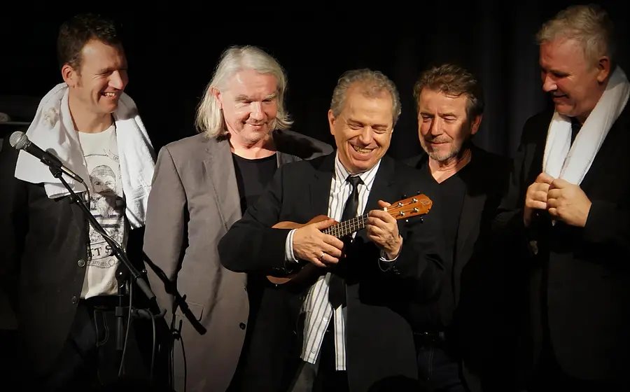 Günther Sigl steht zusammen mit weiteren vier Musikern und hält ein Instrument in der Hand, vor dunklem Hintergrund