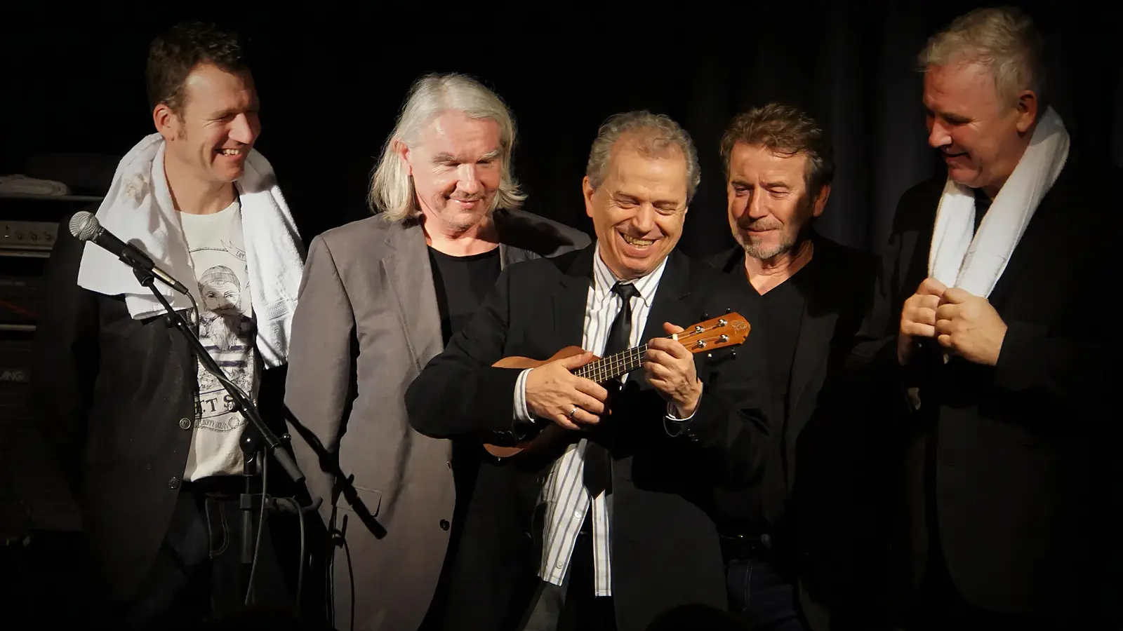 Günther Sigl steht zusammen mit weiteren vier Musikern und hält ein Instrument in der Hand, vor dunklem Hintergrund