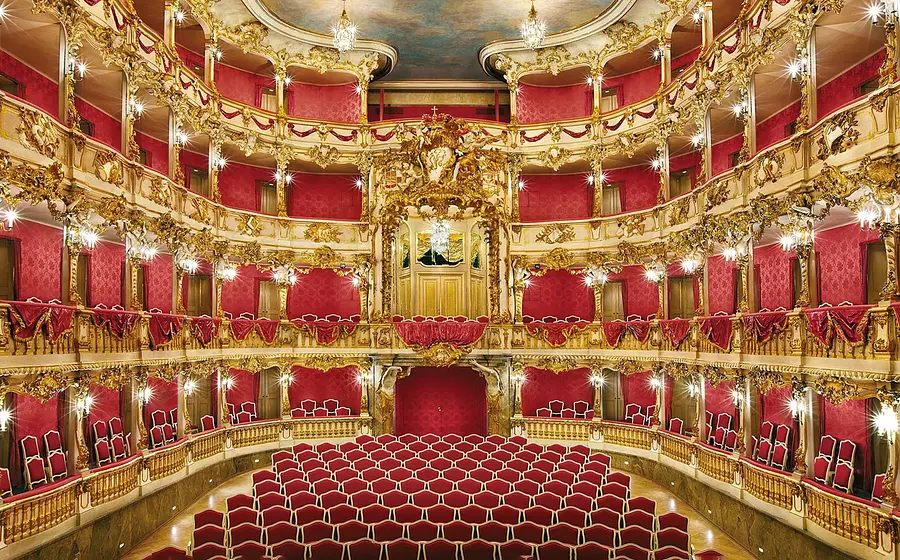 Innenansicht eines prunkvollen barocken Theatersaals mit roten Sitzreihen, goldverzierten Logen, Säulen und mehreren Kronleuchtern, Blick Richtung Bühne mit reich dekorierten Balkonen.
