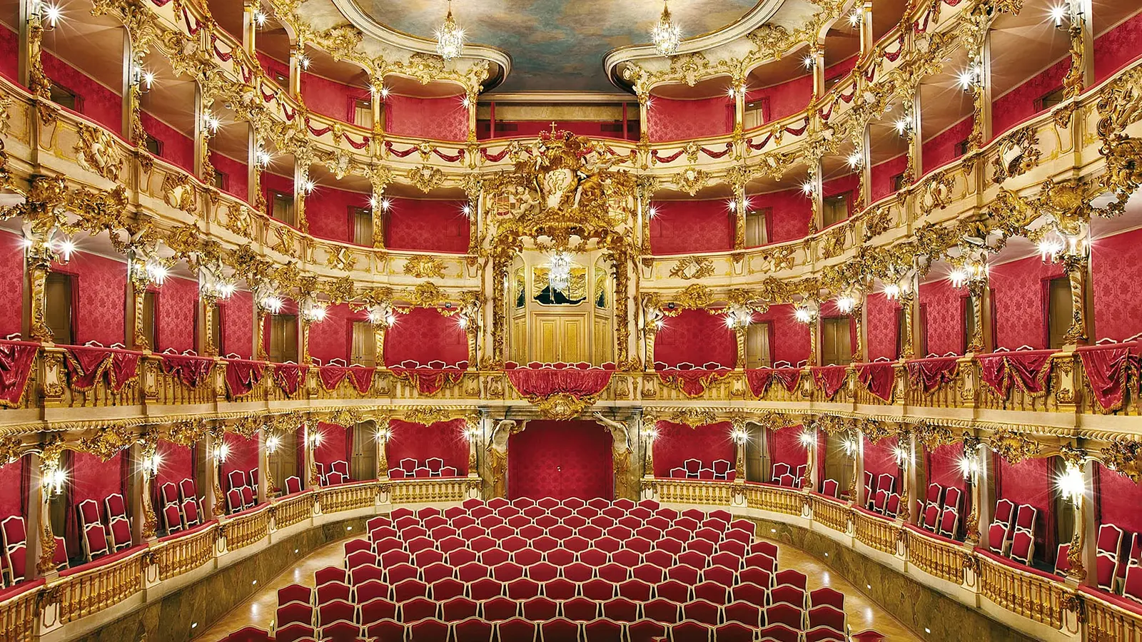 Innenansicht eines prunkvollen barocken Theatersaals mit roten Sitzreihen, goldverzierten Logen, Säulen und mehreren Kronleuchtern, Blick Richtung Bühne mit reich dekorierten Balkonen.