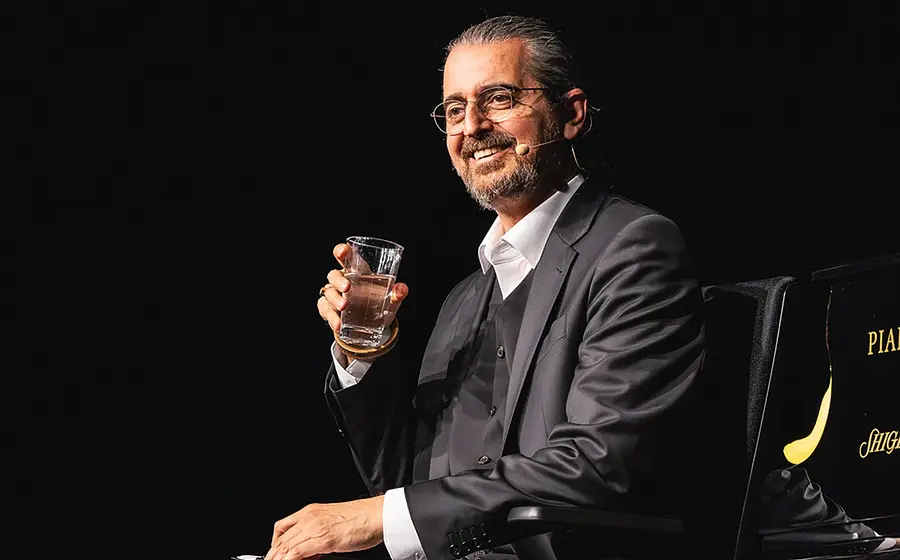 Bühnenfoto von Hagen Rether in dunklem Anzug mit Brille und Headsetmikrofon, sitzend neben einem Flügel und ein Wasserglas haltend, vor schwarzem Bühnenhintergrund.