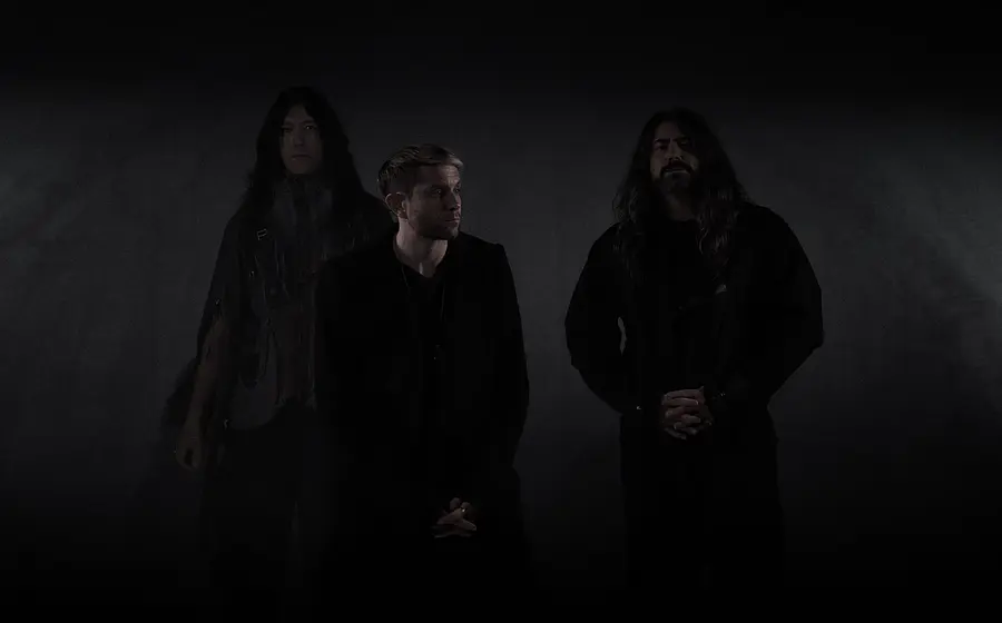 Drei Mitglieder der Band HEALTH stehen vor einem dunklen Hintergrund und blicken ernst; sie tragen schwarze Kleidung und sind minimalistisch beleuchtet, was eine düstere, atmosphärische Stimmung erzeugt.