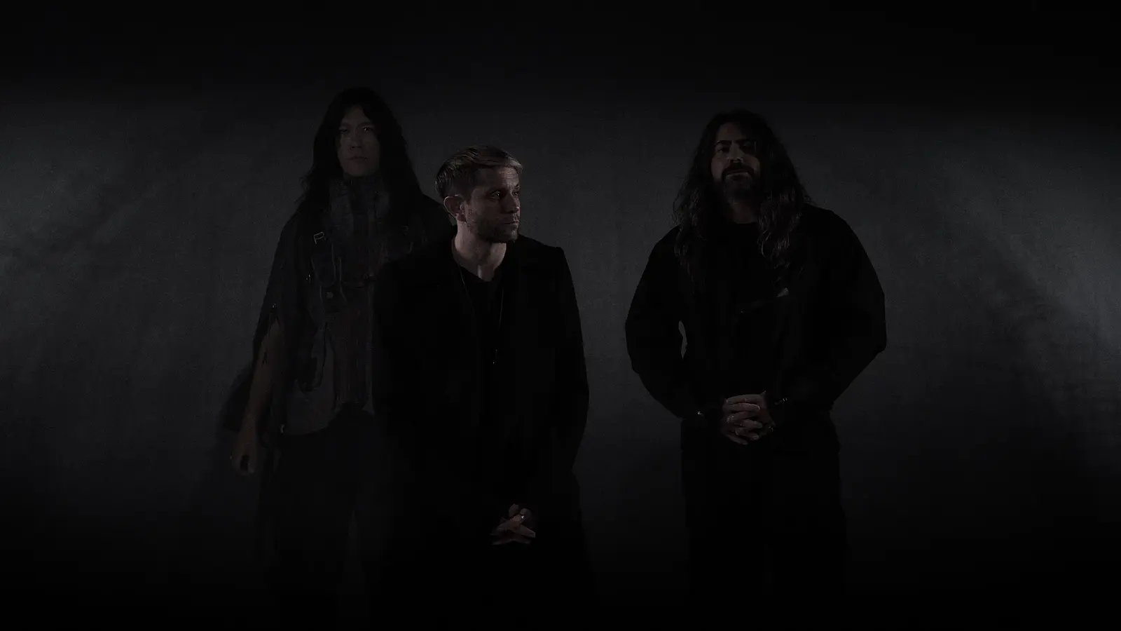 Drei Mitglieder der Band HEALTH stehen vor einem dunklen Hintergrund und blicken ernst; sie tragen schwarze Kleidung und sind minimalistisch beleuchtet, was eine düstere, atmosphärische Stimmung erzeugt.
