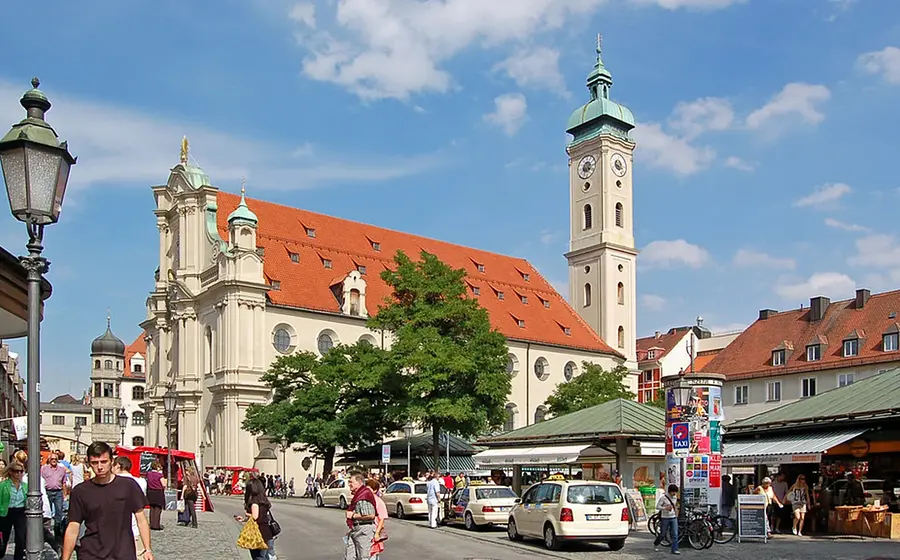Stadtansicht von München am Viktualienmarkt: Blick auf die Heiliggeistkirche mit hellem Kirchturm und rotem Ziegeldach, umgeben von Marktständen, Passantinnen und Passanten sowie parkenden Fahrzeugen; belebte Innenstadt bei blauem Himmel.