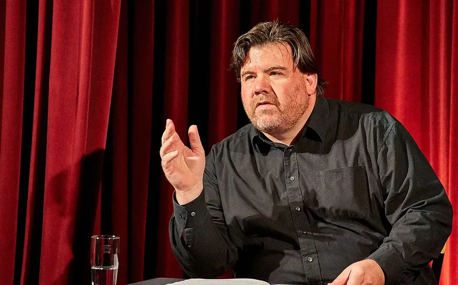 Ein Mann in schwarzem Hemd an einem Tisch sitzend, vor einem roten Vorhang