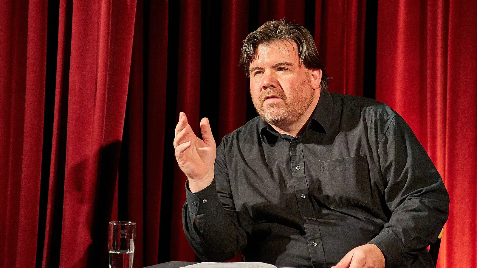 Ein Mann in schwarzem Hemd an einem Tisch sitzend, vor einem roten Vorhang