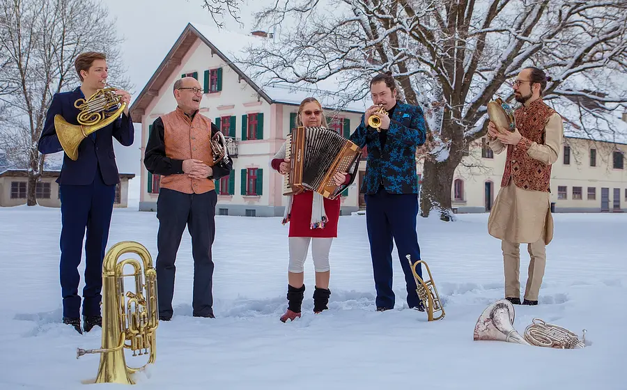Fünf Personen in einer Schneelandschaft mit Blasinstrumenten, im Hintergrund ein traditionelles Bauernhaus