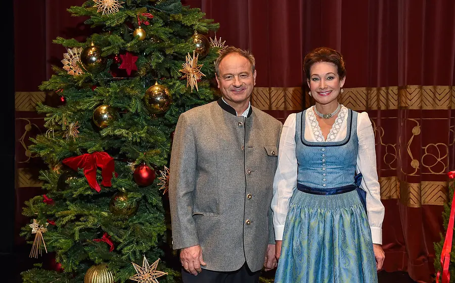 Zwei Personen in Trachtenkleidung vor einem geschmückten Weihnachtsbaum und rotem Bühnenvorhang, eine Person in grauer Trachtenjacke, die andere in blauem Dirndl mit weißer Bluse, nebeneinander stehend.