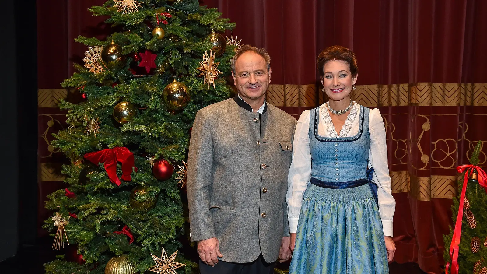 Zwei Personen in Trachtenkleidung vor einem geschmückten Weihnachtsbaum und rotem Bühnenvorhang, eine Person in grauer Trachtenjacke, die andere in blauem Dirndl mit weißer Bluse, nebeneinander stehend.