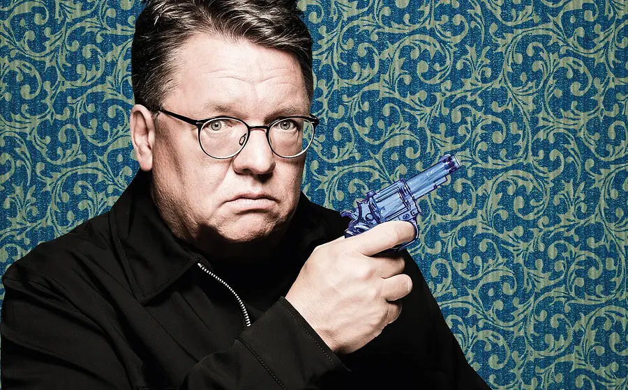 Foto für die Veranstaltung „Helmut Schleich – Live 2026“, mit dem Künstler, der eine Pistole in der Hand hält, vor einem Hintergrund mit blau-grünem Muster.