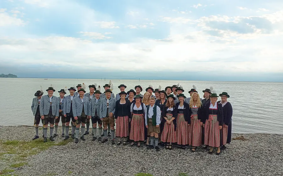 Gruppenfoto der Blaskapelle Unterpfaffenhofen mit Musikerinnen und Musikern in traditioneller Tracht am Ufer eines Sees, vor weiter Wasserfläche und bewölktem Himmel mit Sonnenstrahlen.