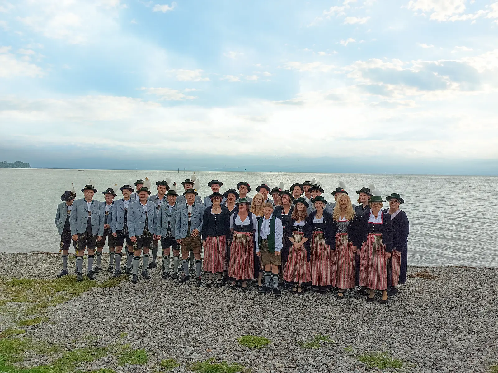 Gruppenfoto der Blaskapelle Unterpfaffenhofen mit Musikerinnen und Musikern in traditioneller Tracht am Ufer eines Sees, vor weiter Wasserfläche und bewölktem Himmel mit Sonnenstrahlen.